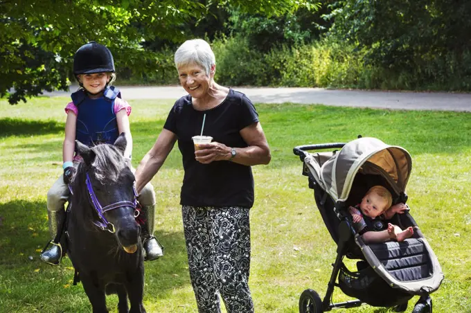 Woman, baby in pushchair and blond girl wearing riding hat sitting on a pony.