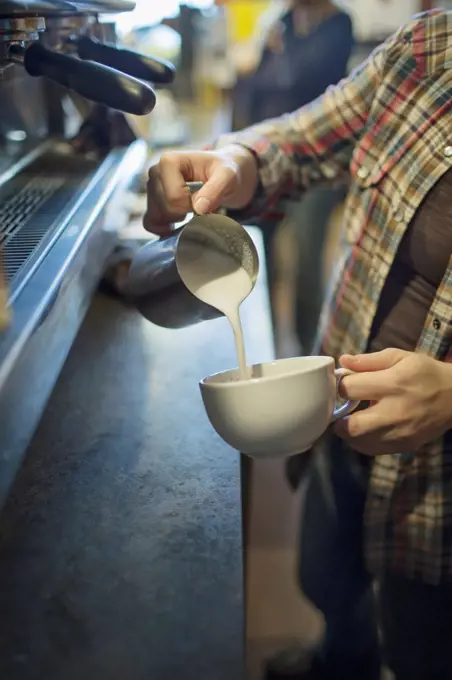 A person, barista, making coffee, and pouring frothed milk from a jug into a cup for a cappuccino. Coffee shop.Kingston, New York, USA