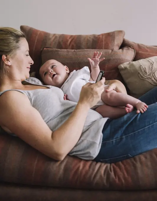 A woman lying on a sofa holding a baby girl, and holding a smart phone in one hand.