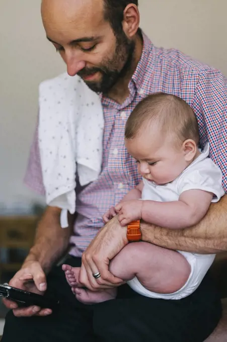 A man holding his small child in the crook of his arm, texting on his smart phone with the other.