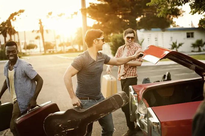 Three young men, friends packing the car with suitcases and a guitar, for a road trip.