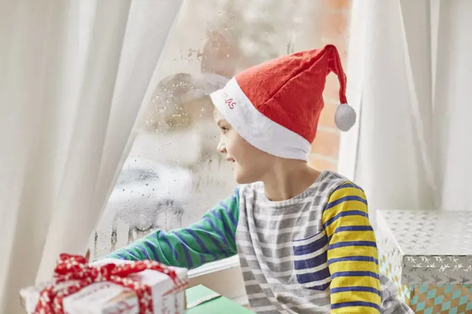 Christmas morning in a family home. A boy in a Santa hat looking out of a bedroom window.