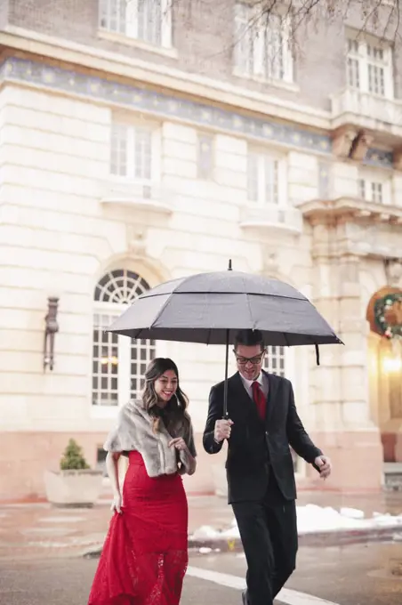 A woman in a long red evening dress with fishtail skirt and a fur stole, and a man in a suit, walking through snow in the city.