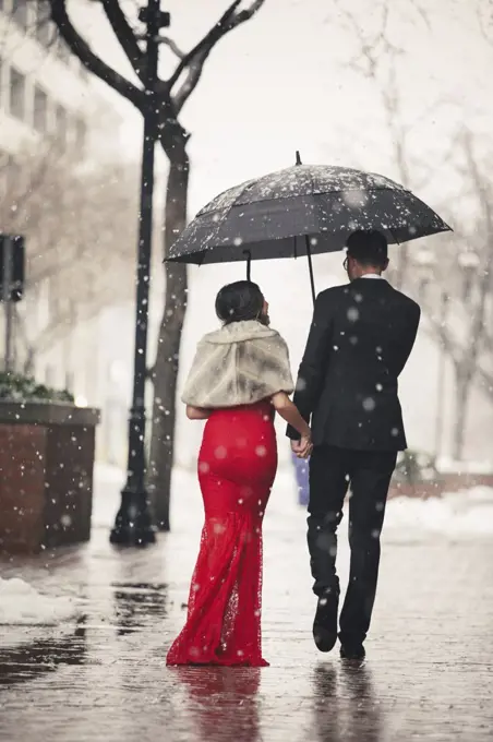 A woman in a long red evening dress with fishtail skirt and a fur stole, and a man in a suit, walking through snow in the city.