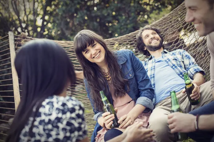 A group of friends lounging in a large hammock in the garden having a beer.