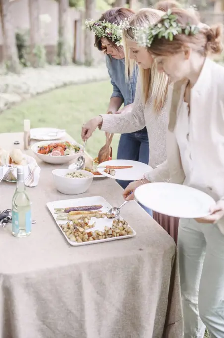 Women with flower wreath in their hair at a garden party, choosing food from a buffet.