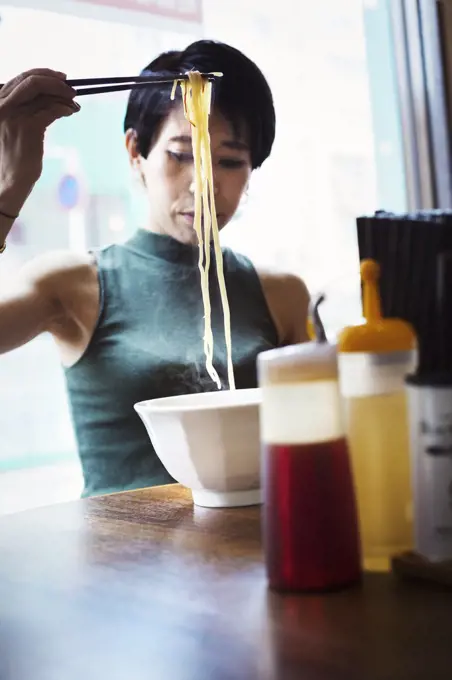 A ramen noodle cafe in a city.  A woman seated eating a ramen noodle dish using chopsticks.