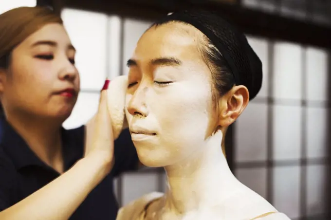 A modern geisha or maiko woman being prepared in traditional fashion, with white face makeup.