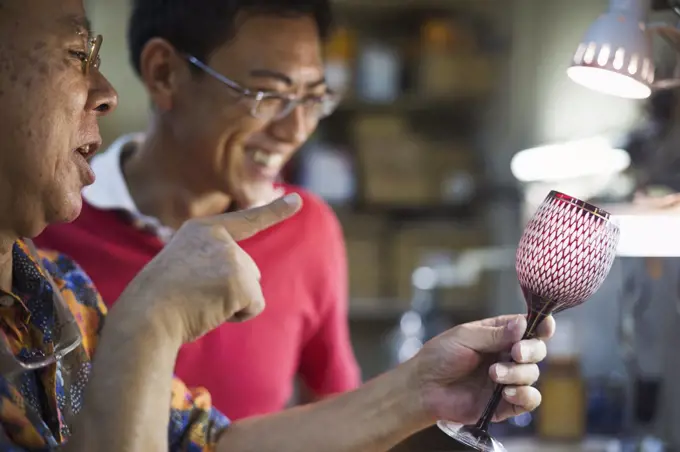 Two people, a father and son at work in a glass maker's studio workshop, inspecting a red cut glass wine glass.