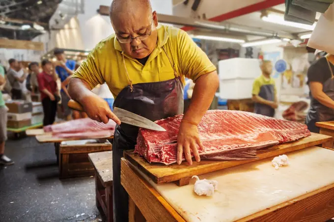 A traditional fresh fish market in Tokyo. A fishmonger working filleting a large fish on a slab. People in the background.