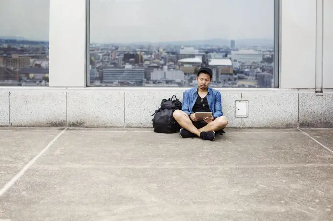 A man sitting on the floor using his smart phone, in front of a viewing point window on to a city.