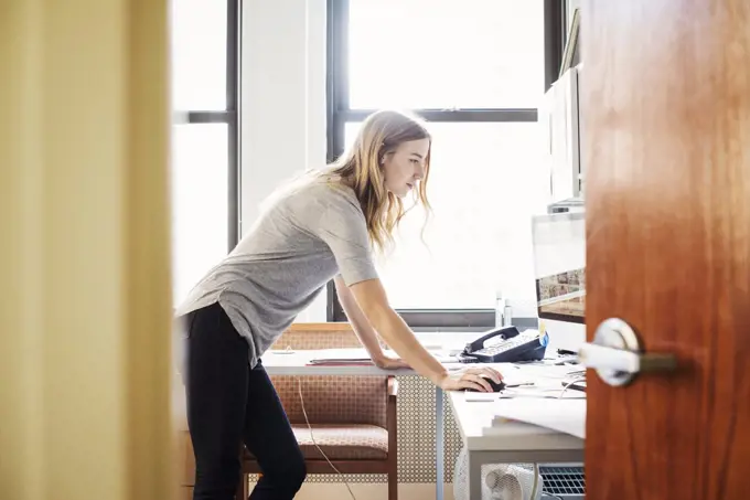 A young woman in an office standing over a desk and working at a computer.