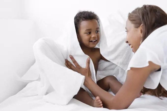 Women wrapping young boy sitting on a bed in white towel, looking at each other, smiling.