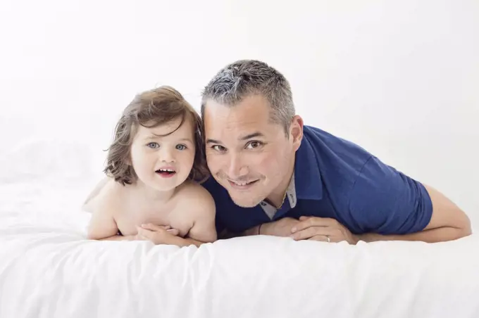 Man and young girl with brown hair lying side by side on a bed, smiling at camera.