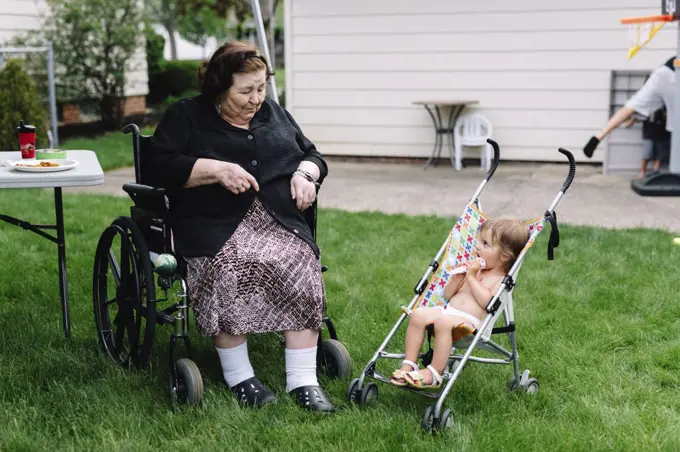 Smiling senior women sitting in a wheelchair in a garden beside bare chested young girl sitting in stroller.