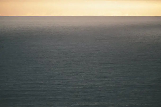 View from the land over the ocean, to the horizon at dusk. Light orange coloured sky and dark water.