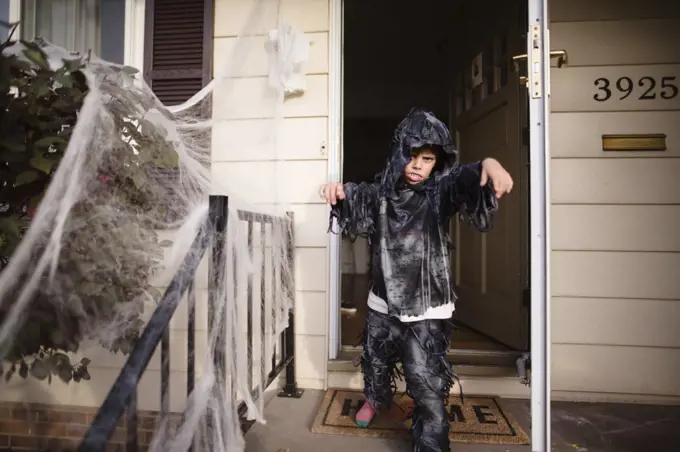 Boy wearing hooded Halloween costume standing outside front door of house, banister decorated with white netting.