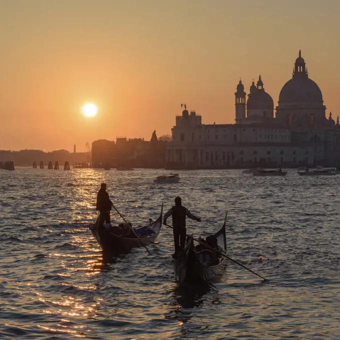 Two gondoliers on the Canale Grande in Venice, Italy, at sunrise, with the dome of Santa Maria della Salute in the distance.