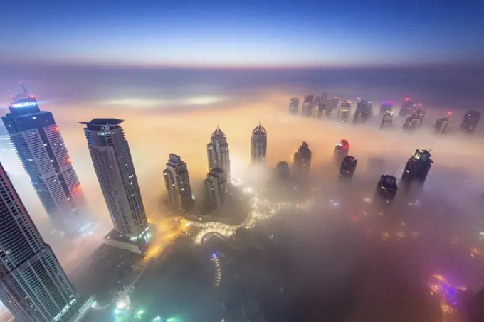 Aerial view of cityscape with illuminated skyscrapers above the clouds in Dubai, United Arab Emirates at dusk.