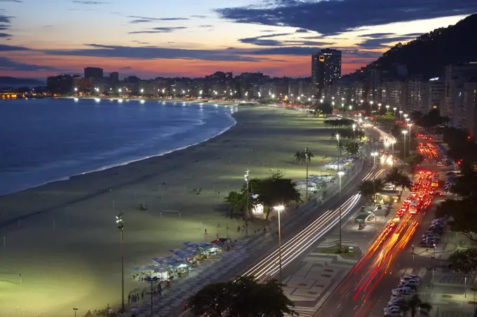 High angle view of long sandy beach lined by illuminated tall buildings, light trail of cars on avenue.