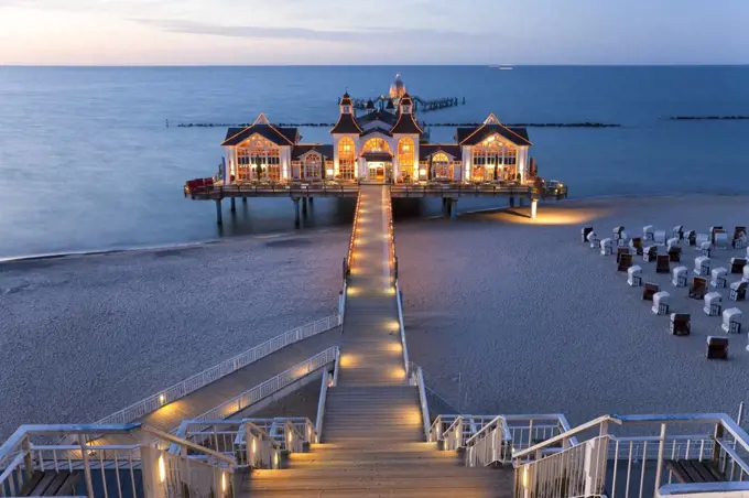 Wooden stairway and walkway leading across sandy beach towards illuminated building on a pier.