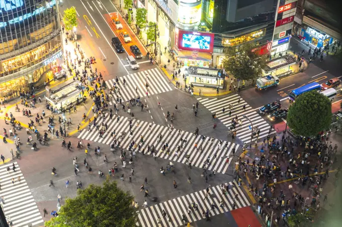 High angle view of urban street lined with illuminated buildings, pedestrians walking on pedestrian crossings.