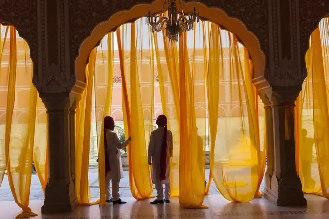 Two men wearing traditional clothing and turbans standing under arch decorated with translucent orange curtains.