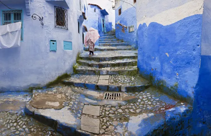 Rear view of person climbing cobblestone steps lined with blue washed traditional North African houses.