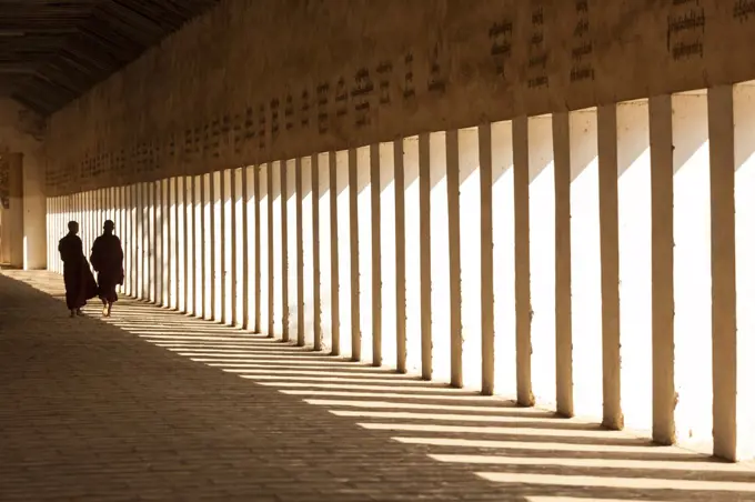 Rear view of two monks walking along sunlit colonnade of pagoda.