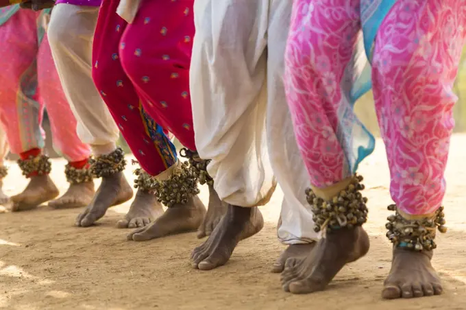 Low section view of barefoot dancers in a row on a street, wearing pink trousers and ankle bells.