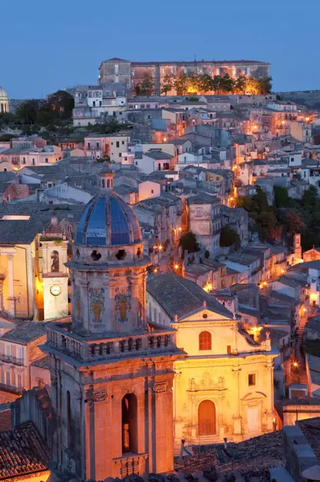 High angle view over illuminated rooftops of traditional houses in a Mediterranean city at dusk, church cupola in the foreground.