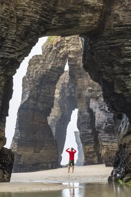 Rear view of man wearing red top standing underneath natural rock arch on sandy beach.