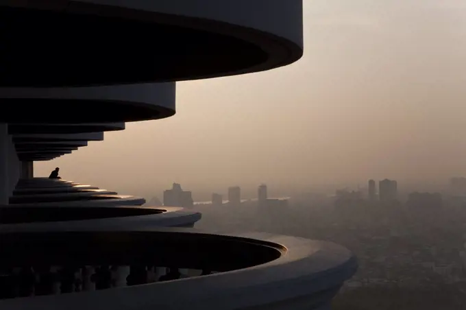 Man standing on curved balcony on skyscraper, looking over misty cityscape.