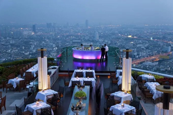 High angle view of rooftop restaurant on a skyscraper, illuminated cityscape in the distance.