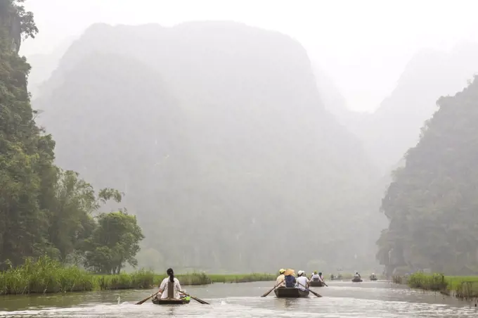 Groups of people in rowboats on a river, mountains in the background.