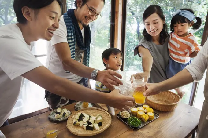 Two men, woman holding young girl and boy gathered around a table with food, holding drinking glasses, toasting.