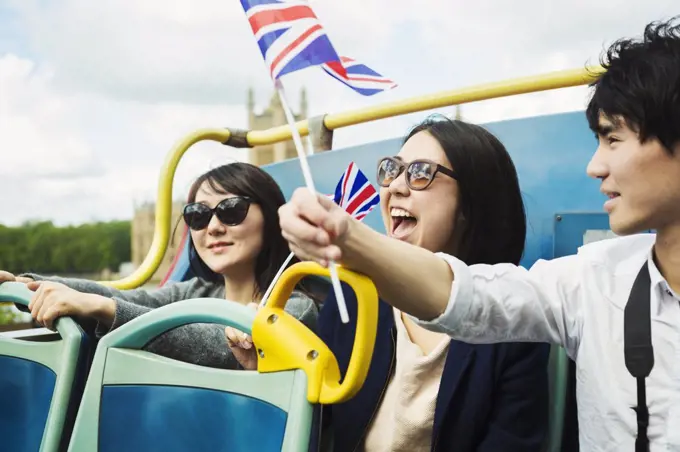 Smiling man waving Union Jack flag and two women with black hair sitting on the top of an open Double-Decker bus.