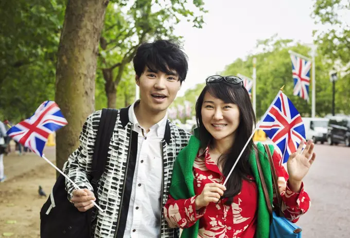 Smiling man and woman with black hair standing on the side of a tree-lined urban road, holding small Union Jack flags, looking at camera.