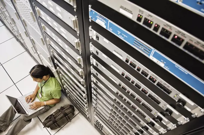 Computer technician working on a lap top computer while doing diagnostic work on a server in a larger computer server room.