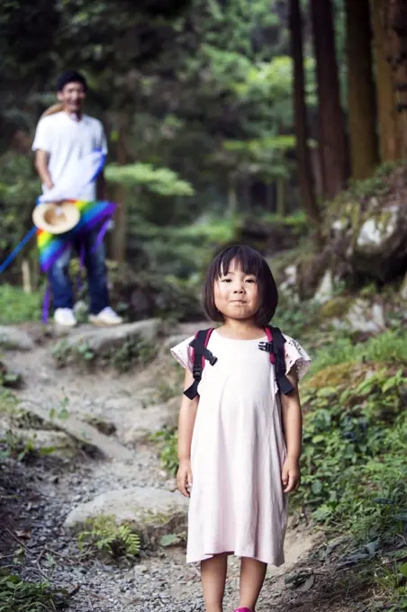 Japanese girl wearing pale pink sun dress and carrying backpack standing in a forest, man in the background.