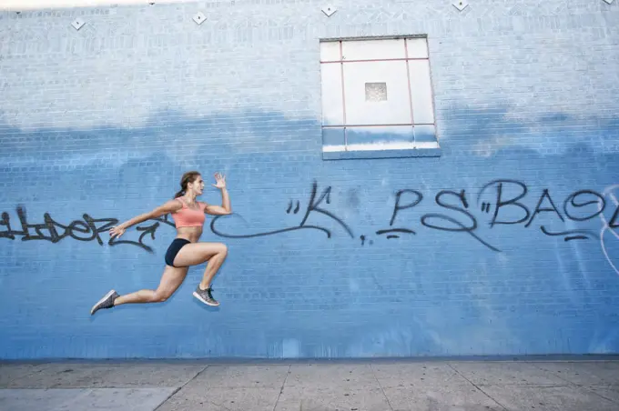 Female athlete running along sidewalk past blue building covered in graffiti.