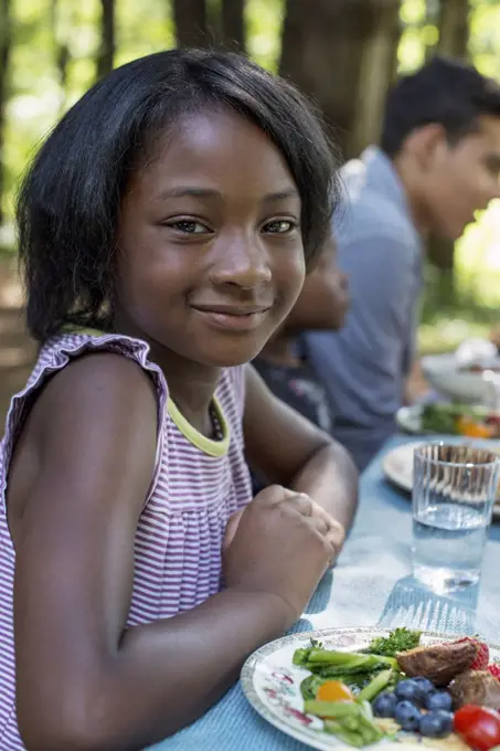 A family picnic meal in the shade of tall trees. A young girl seated at the table. Woodstock, New York, USA