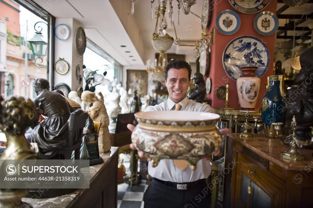 Portrait of a smiling mid-adult businessman holding an antique bowl while standing inside an antique store.