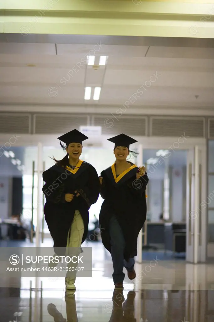 Two teenage girls wearing graduation regalia running down the corridor.