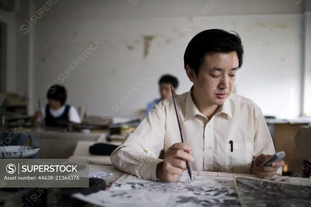 Mid-adult man painting and texting in an art classroom.