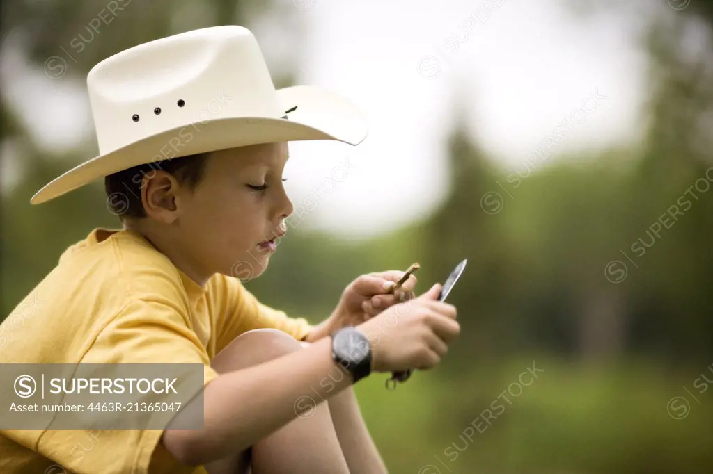 Young boy shaping a twig with a pocketknife while wearing a hat.