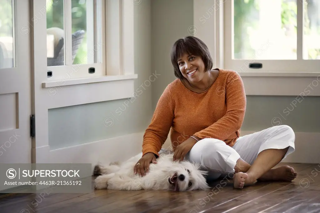 Middle-aged woman smiles and relaxes by petting her dog on the floor.