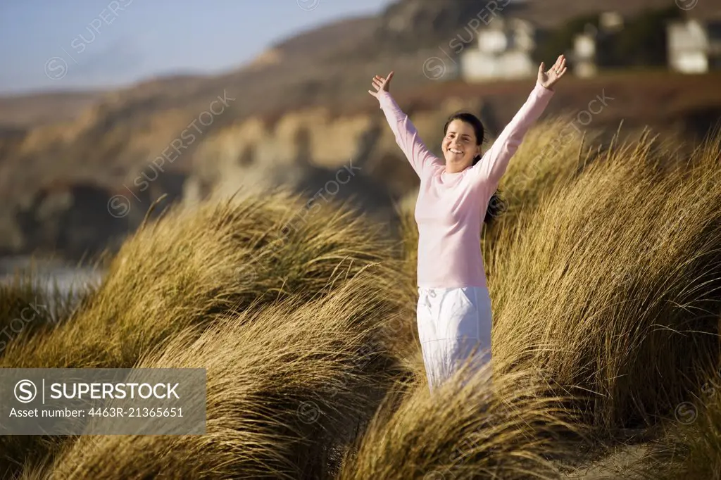 Smiling mid-adult woman standing on a sand dune with her arms raised.