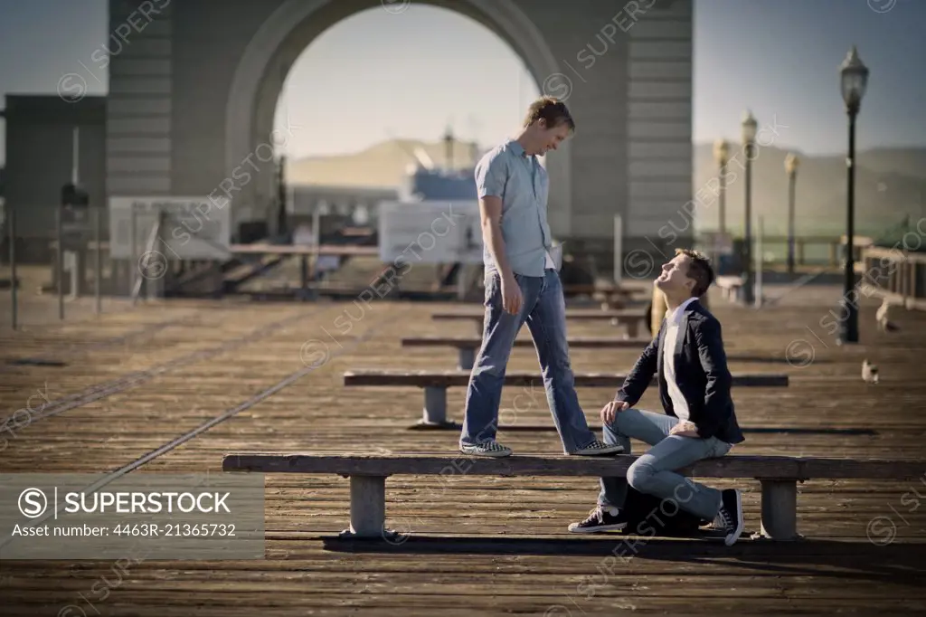 Mid-adult man sitting on a bench looking up at another mid-adult man outside.