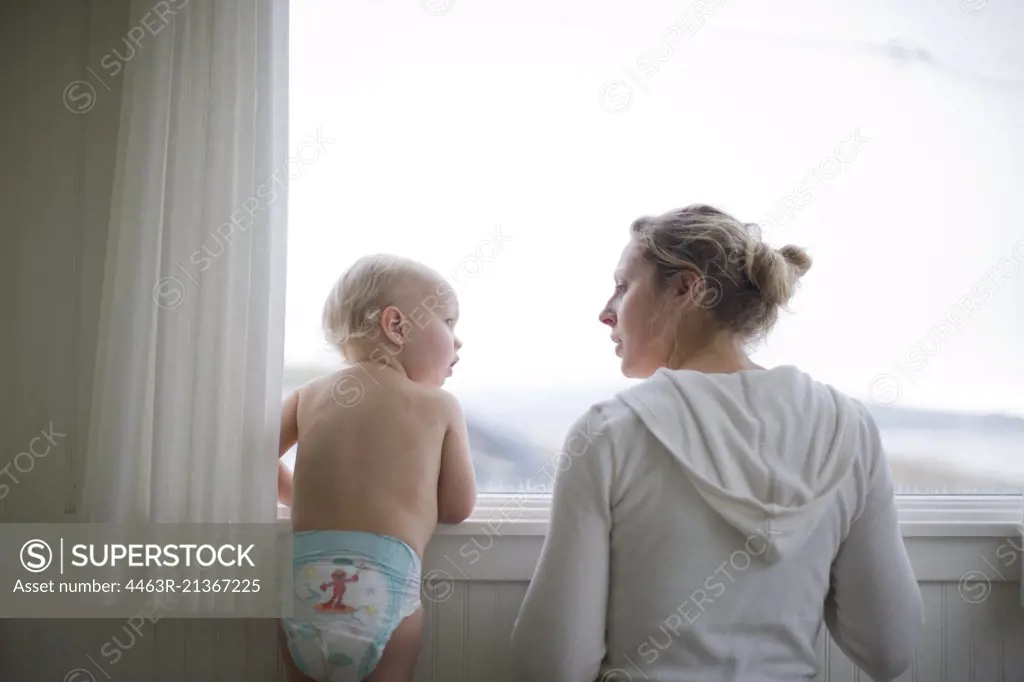 Toddler looking at his young adult mother as they look through a window.